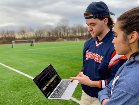 Two individuals on a soccer field, intently observing a laptop screen together.